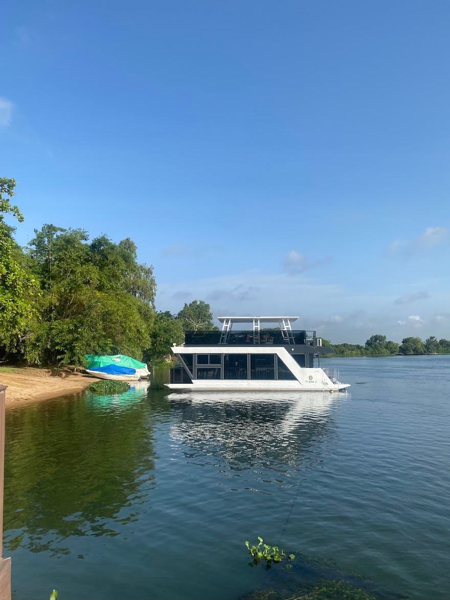 A glass-enclosed boat on the calm river beside the tree-lined bank.